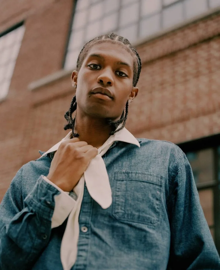 Young man in denim shirt with white collar standing in front of brick building