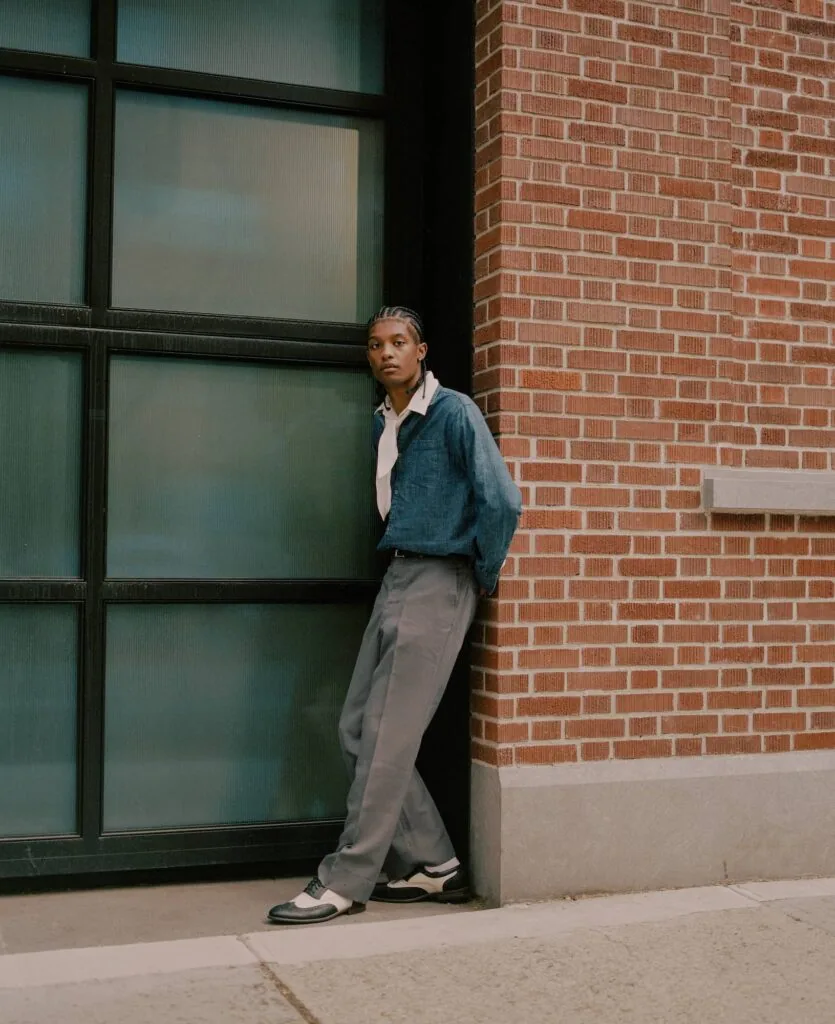 Person in denim jacket and gray pants standing against brick building with metal-framed window