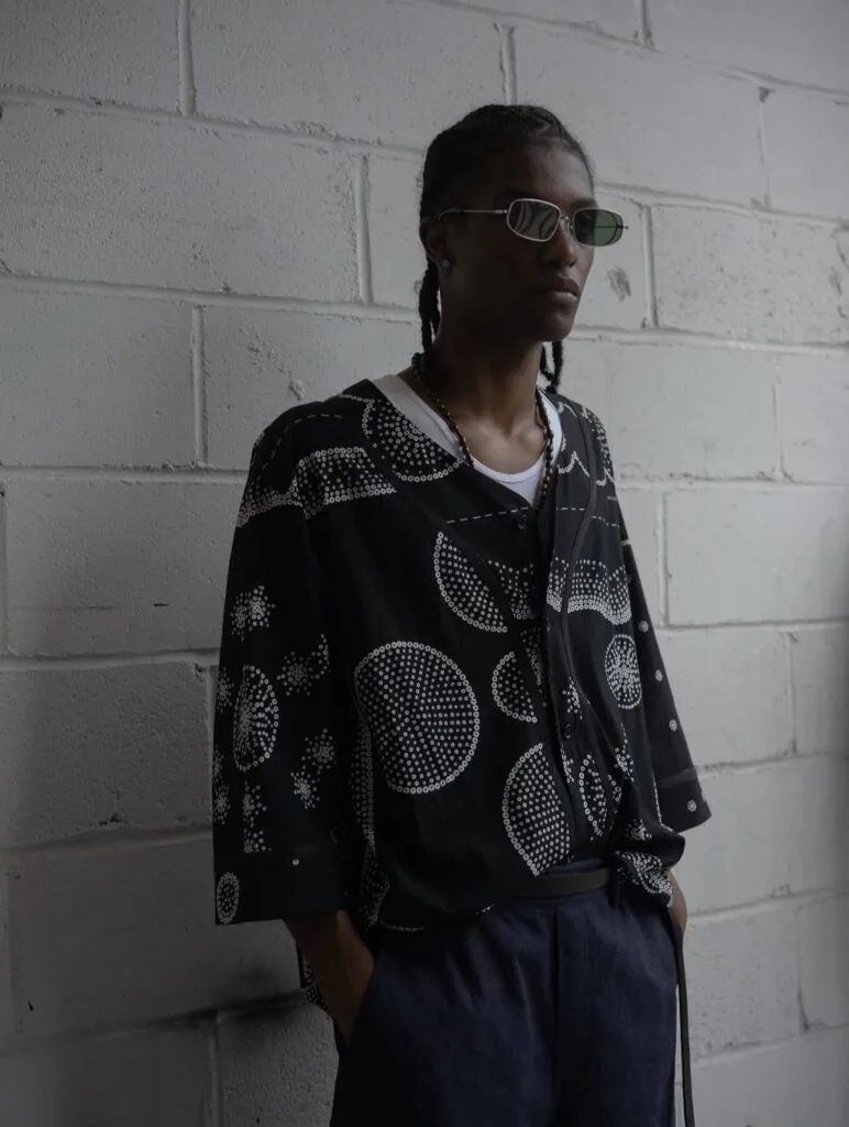 Person wearing sunglasses and a black patterned shirt with circular dot designs standing against a white brick wall