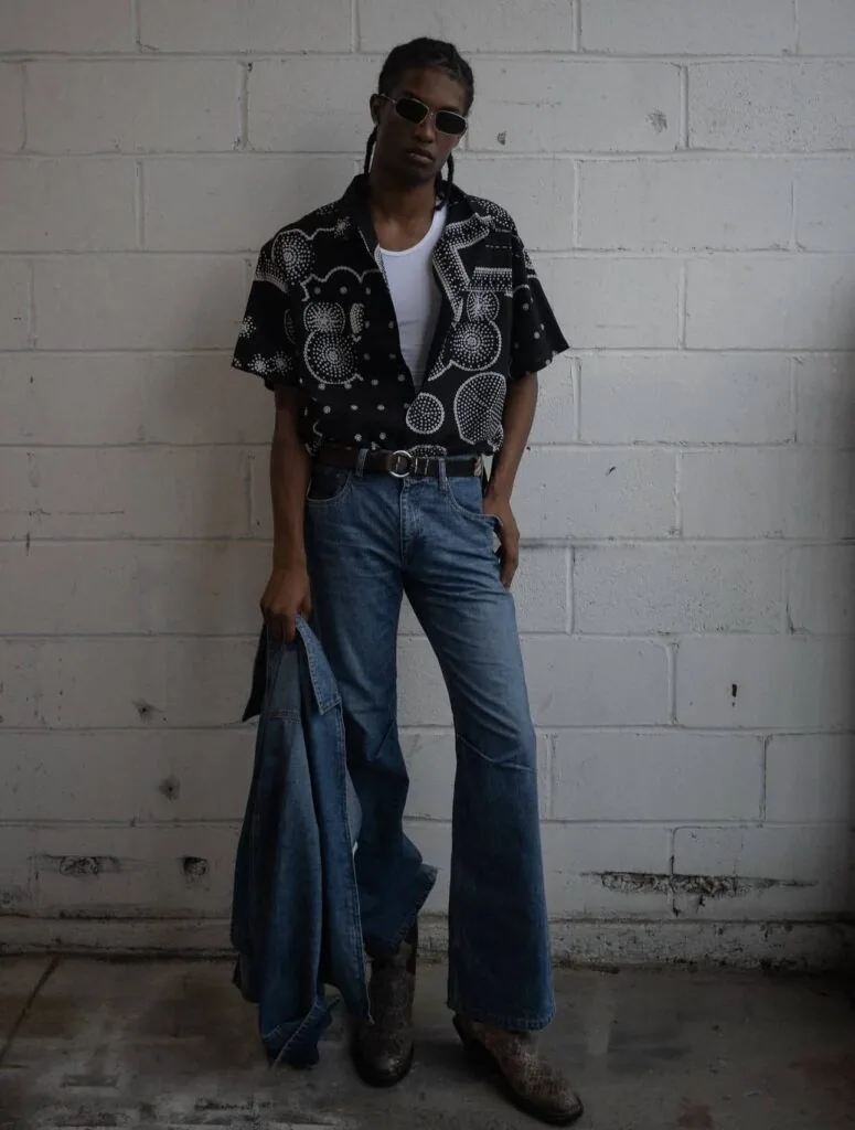 Person wearing sunglasses, patterned black shirt, white top, and blue jeans standing against white brick wall