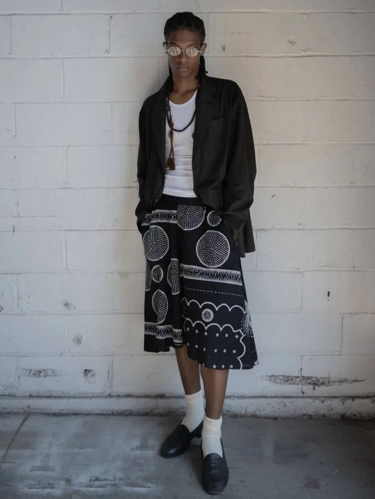Person wearing glasses, black cardigan, white tank top, and black skirt with geometric white patterns, standing against white brick wall