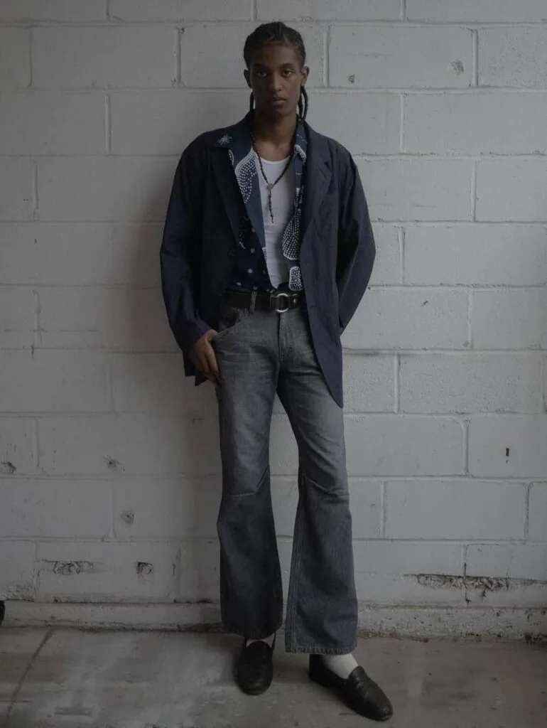 Person wearing navy cardigan, white shirt, gray flare jeans, and black belt standing against white brick wall