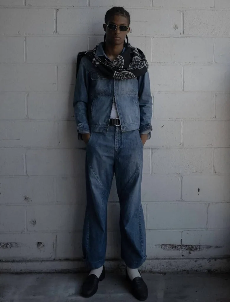 Person wearing denim jacket and wide-leg jeans with patterned scarf against white brick wall