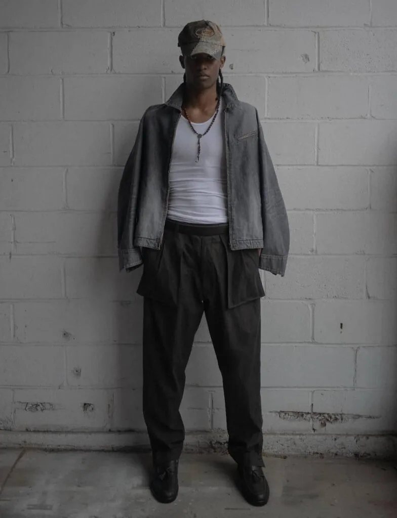 Person wearing denim jacket, white tank top, and black pants standing against white brick wall