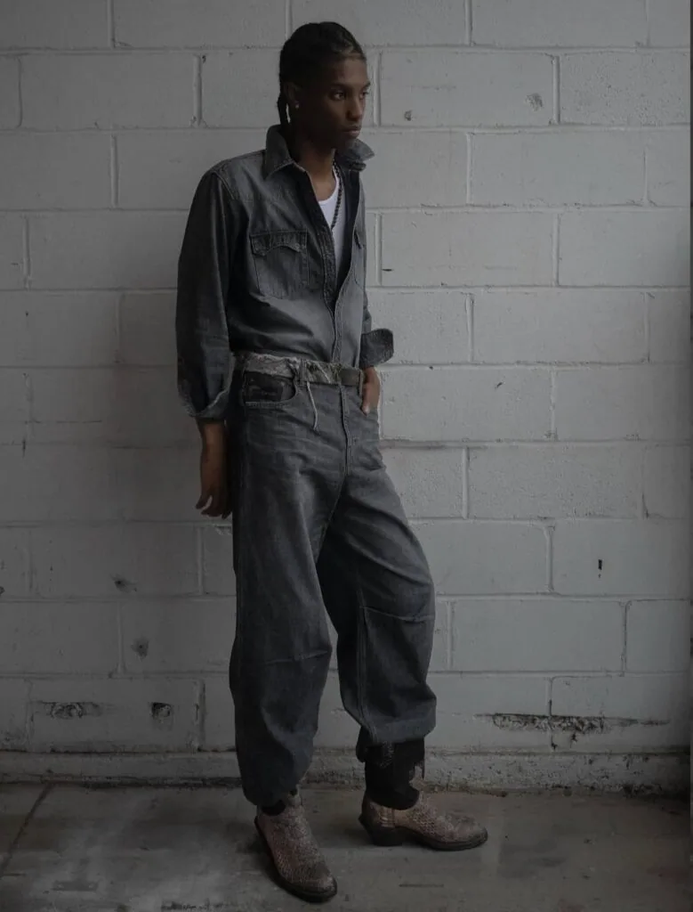 Person wearing denim shirt and jeans standing against white brick wall