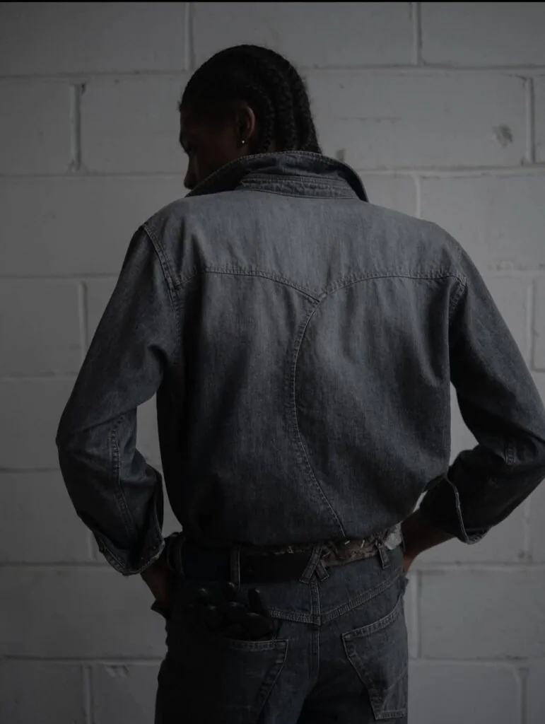 Person wearing dark denim jacket and jeans standing against white brick wall, shown from behind