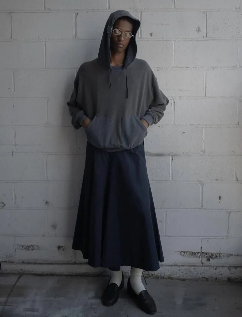Person wearing gray hoodie, dark skirt, white socks and black shoes standing against white brick wall