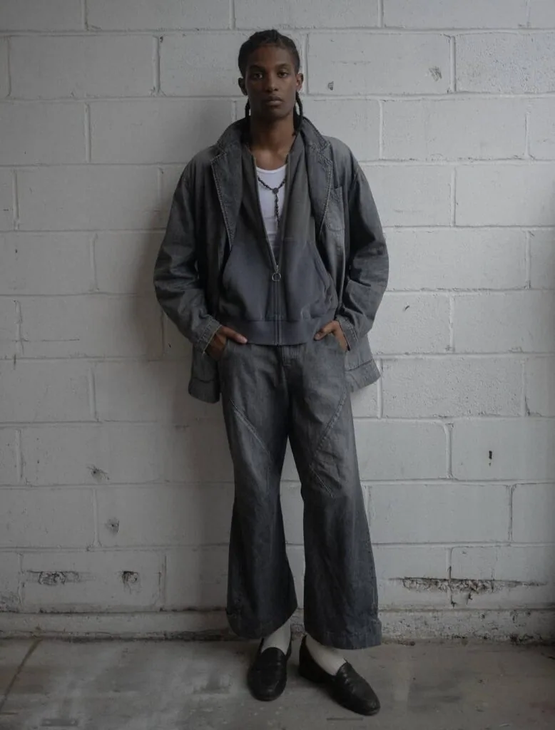 Person standing against white brick wall wearing gray denim jacket and pants with white undershirt