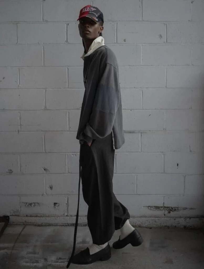 Person in dark oversized clothing and red cap standing against white brick wall holding a cane