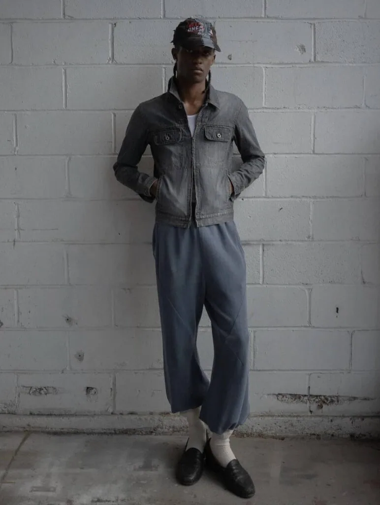 Person wearing denim jacket, navy pants, white socks, and black shoes standing against white brick wall
