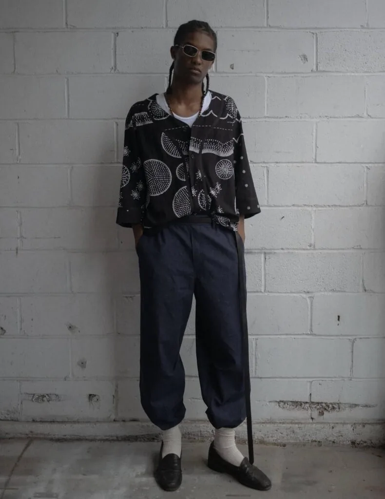 Person wearing sunglasses and a black patterned shirt with white collar over navy pants and loafers, standing against a white brick wall.